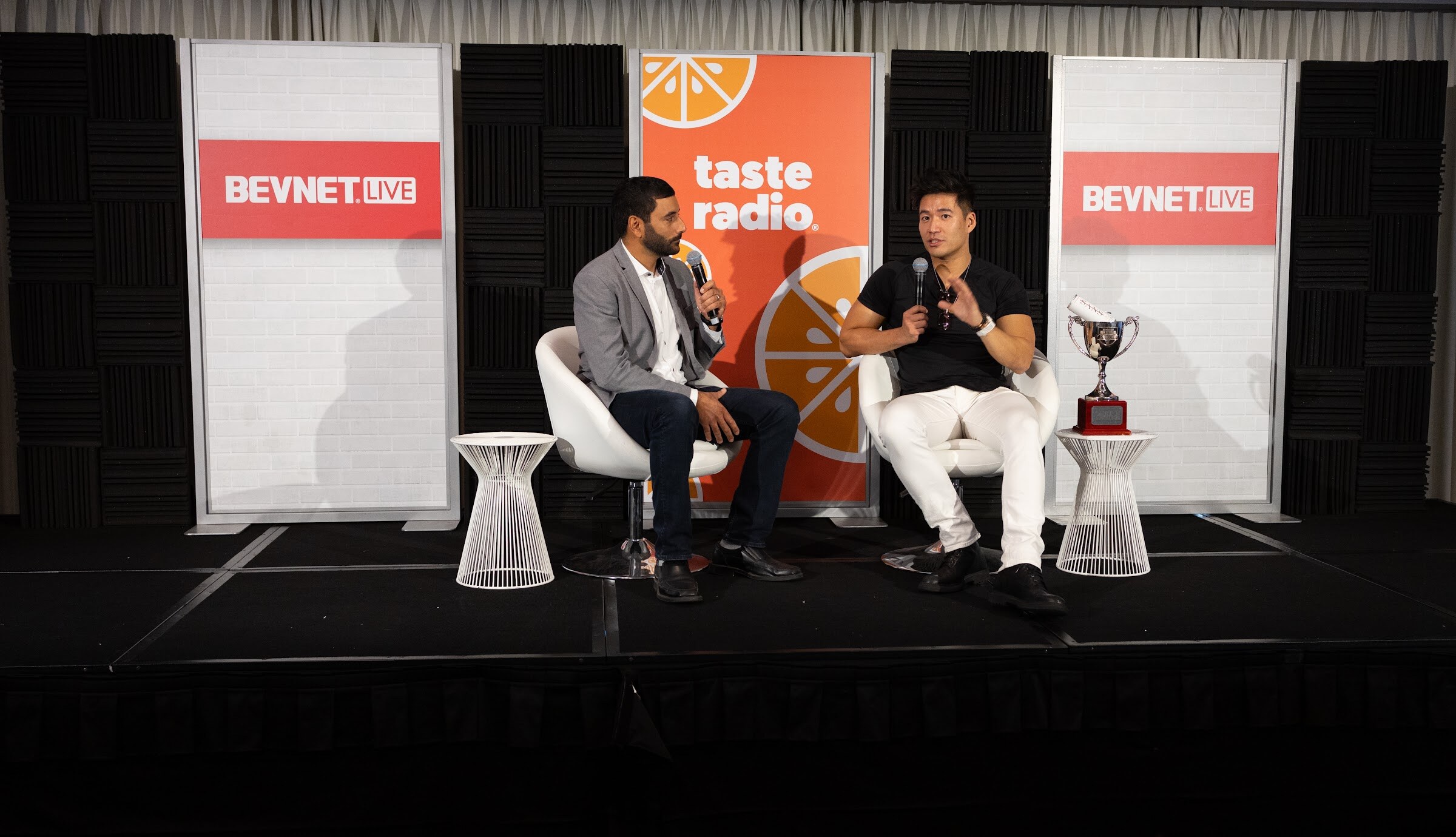 Two men sit in white chairs between BevNET Live signs and a Taste Radio banner, one in a gray blazer and one in a black t-shirt, with a trophy on the table beside them.