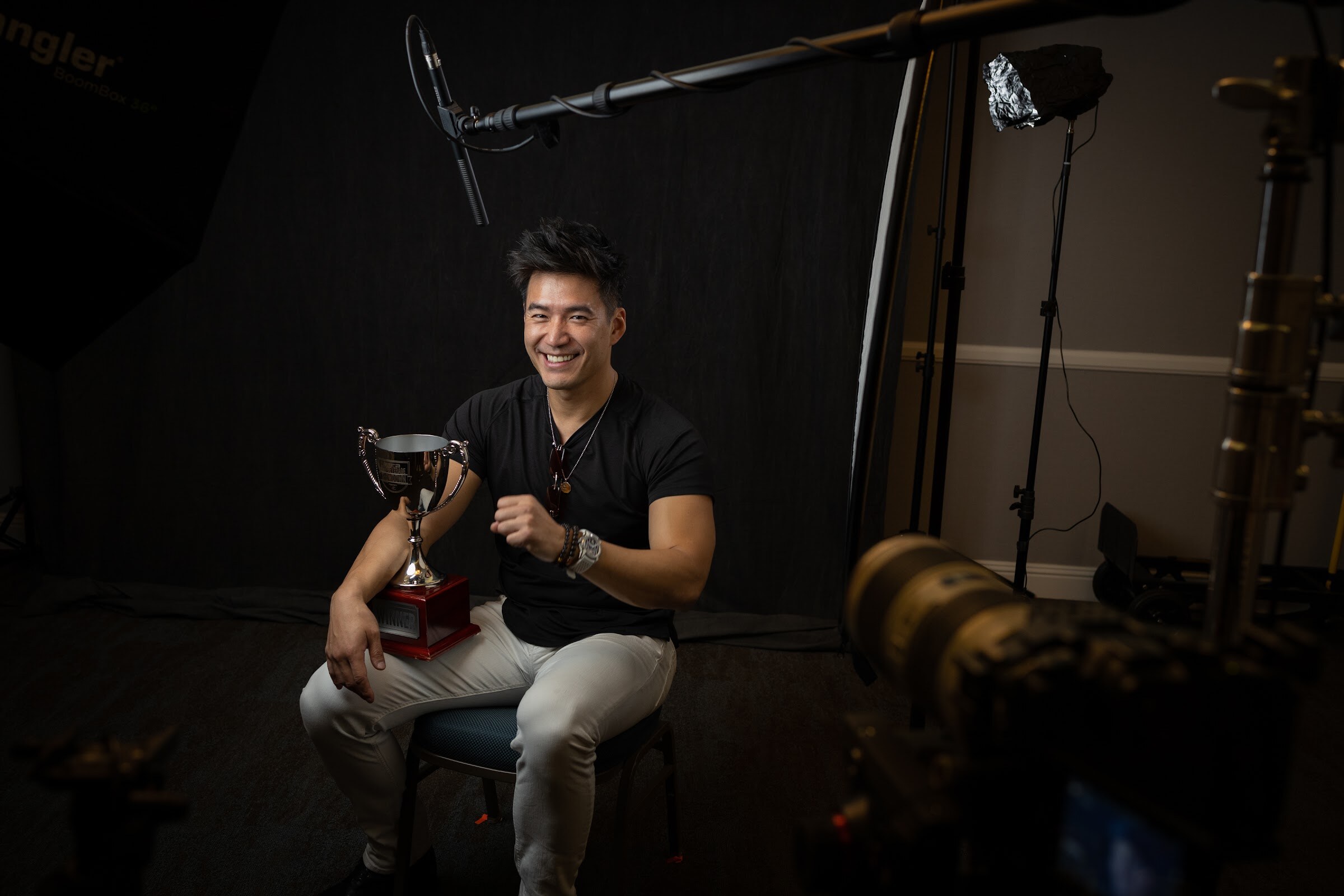 A man in a black t-shirt and gray pants sits on a stool holding a silver trophy cup in a dark room with a boom microphone and lighting stands.