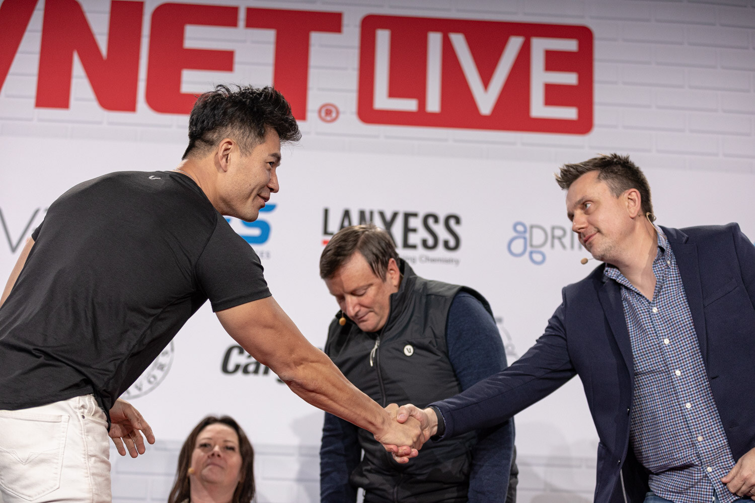 A man in a black t-shirt shakes hands with a man in a navy blazer while two seated judges look on.