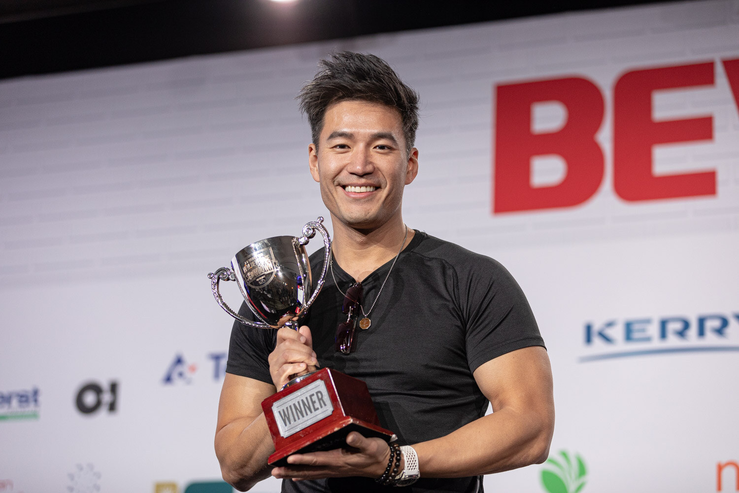 A smiling man in a black t-shirt holds a silver cup trophy with a red base labeled Winner in front of a BevNET Live sign.
