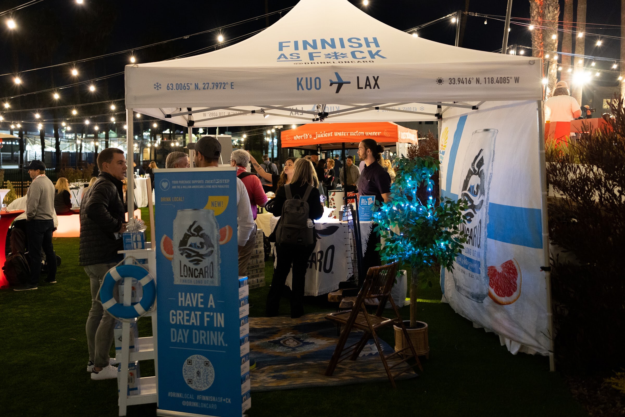A white canopy tent reading Finnish as F*ck KUO to LAX with a Long Drink banner and attendees under string lights on a lawn at night.