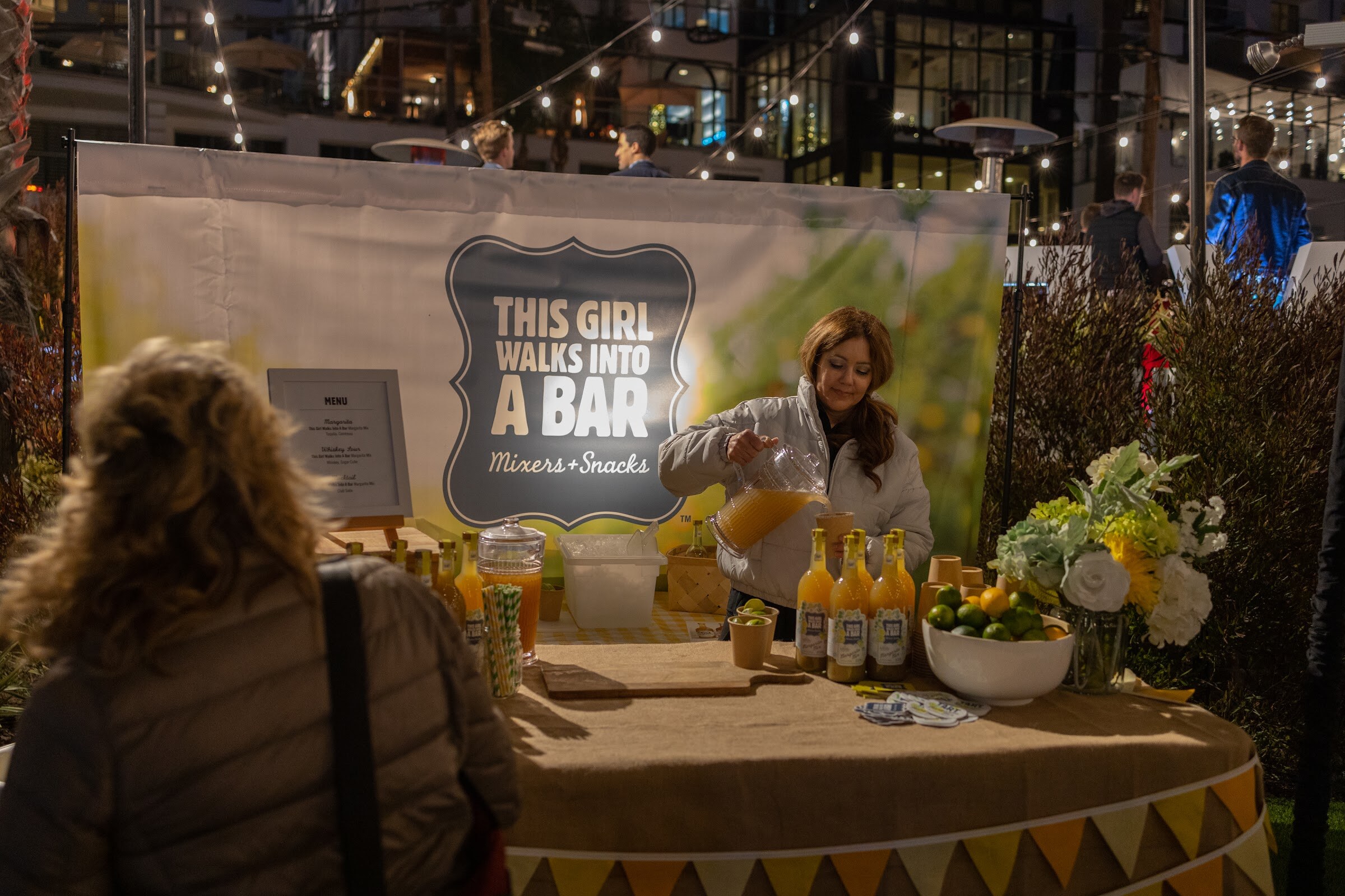 A woman in a white jacket pours juice from a pitcher at a burlap-draped table with bottled mixers and limes in front of a This Girl Walks Into a Bar sign.