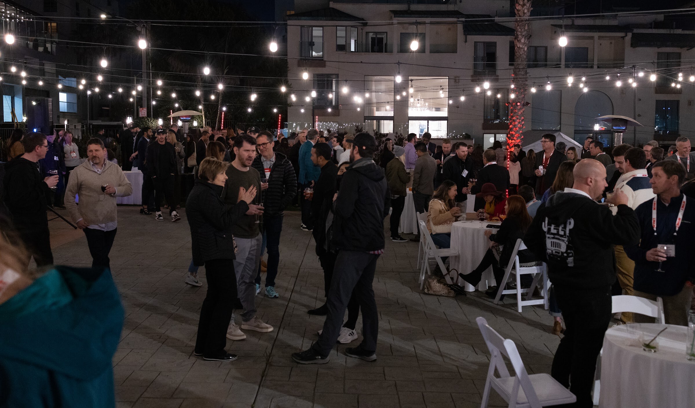 A crowd of attendees stands and talks on a paved plaza at night under strung lights, with white chairs and round tables to the side.