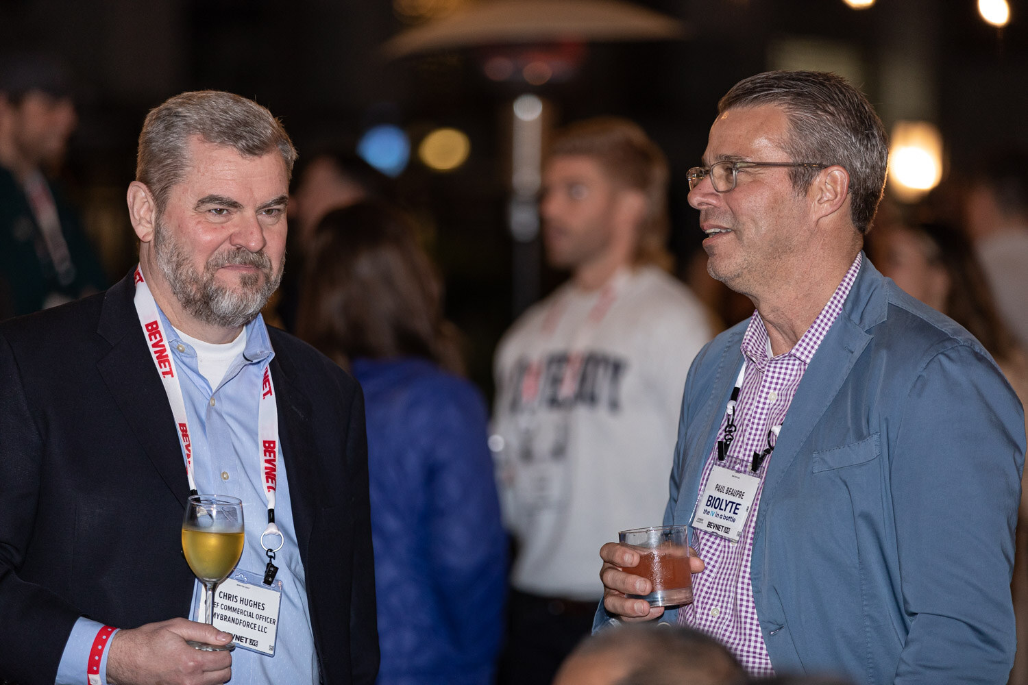 A bearded man in a black blazer holding a glass of white wine and a man in a blue blazer holding a cocktail look off to the side at a crowded outdoor party.