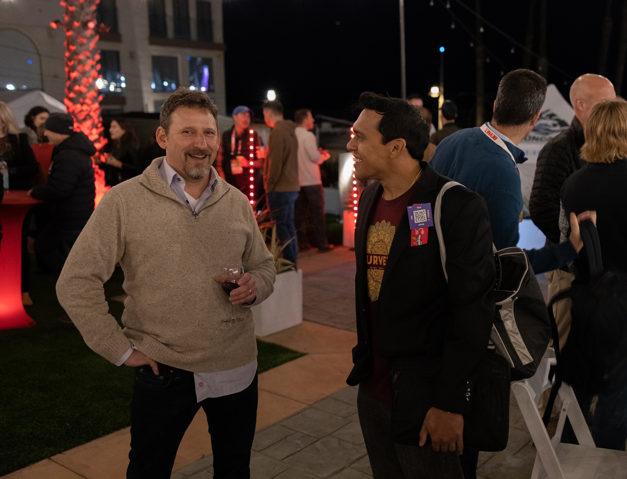 A man in a tan sweater holding a wine glass laughs with a man in a black jacket on a lit lawn.