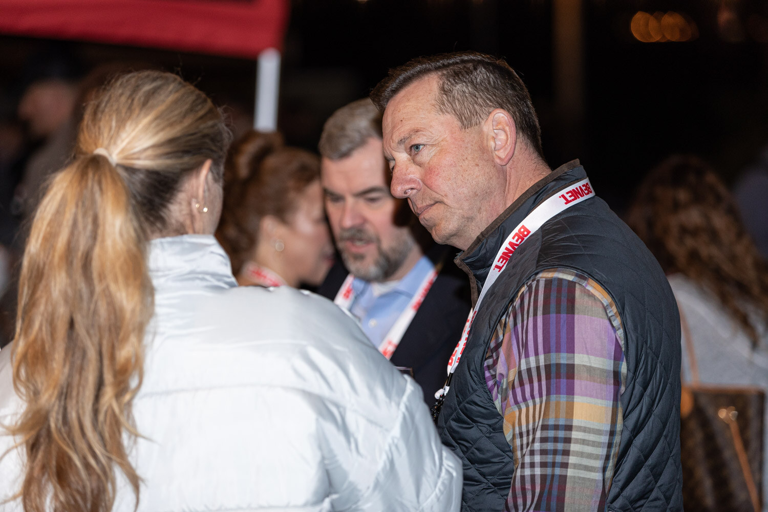 A man in a plaid vest with a BevNET lanyard listens to a woman with a ponytail in a white puffer jacket, with others behind them.