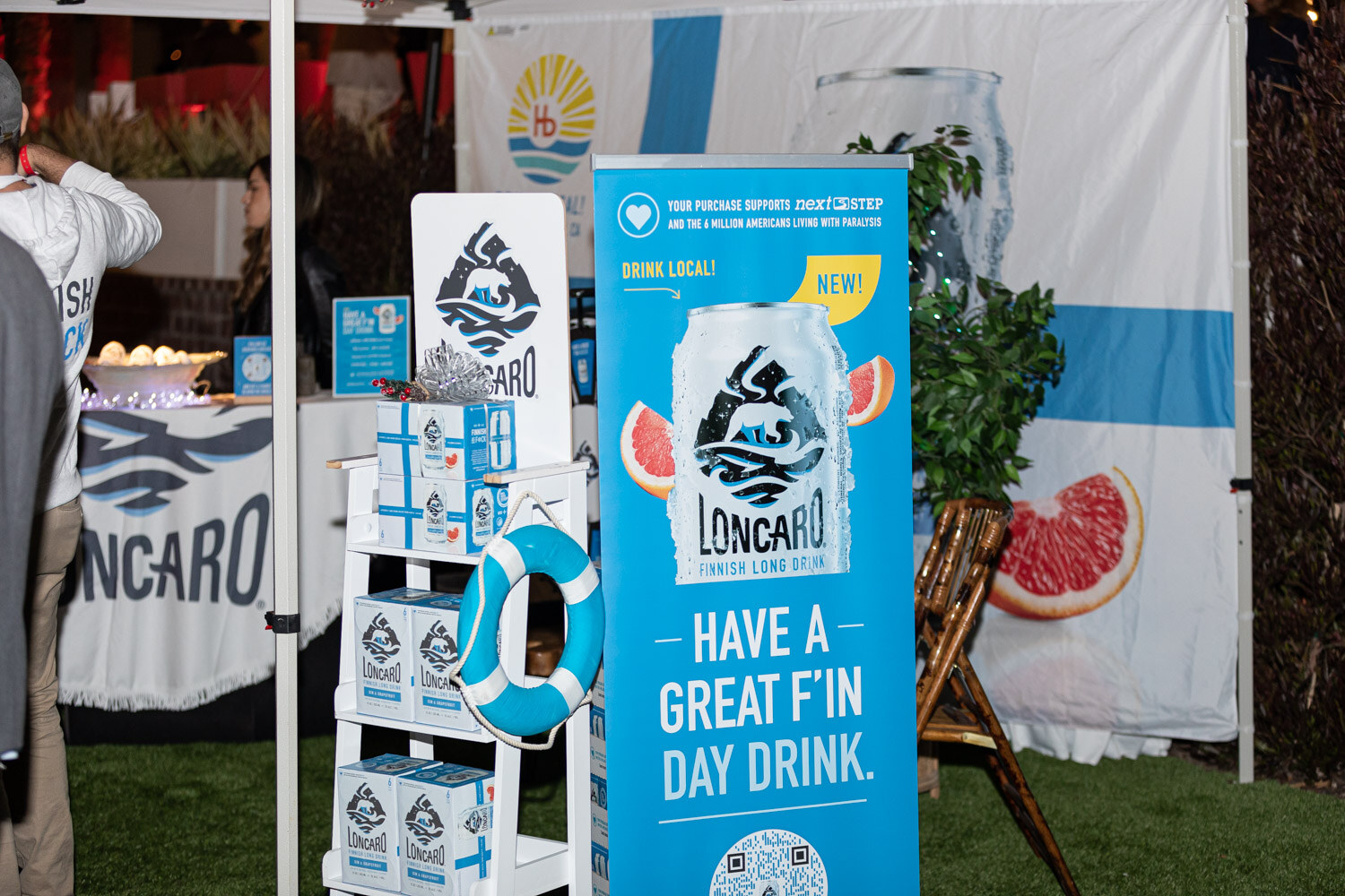 A tent on the lawn with a blue banner reading Have a great f'in day drink with a can, a life ring, a grapefruit slice graphic and a Finnish flag backdrop.