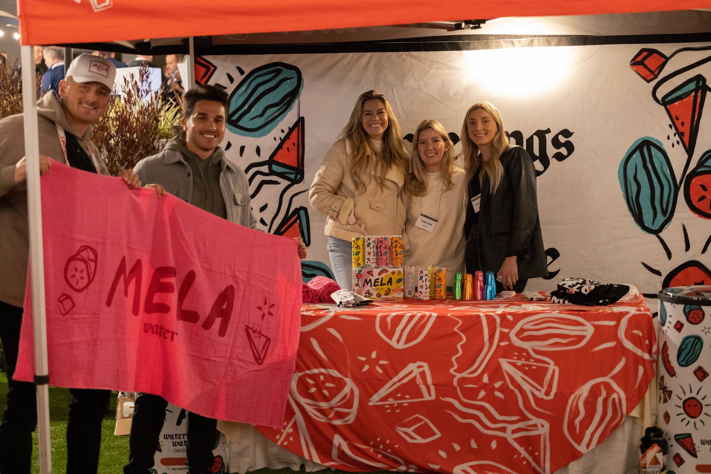 Five people in jackets stand behind a red-patterned table with Mela cans while two of them hold a pink towel reading Mela water.