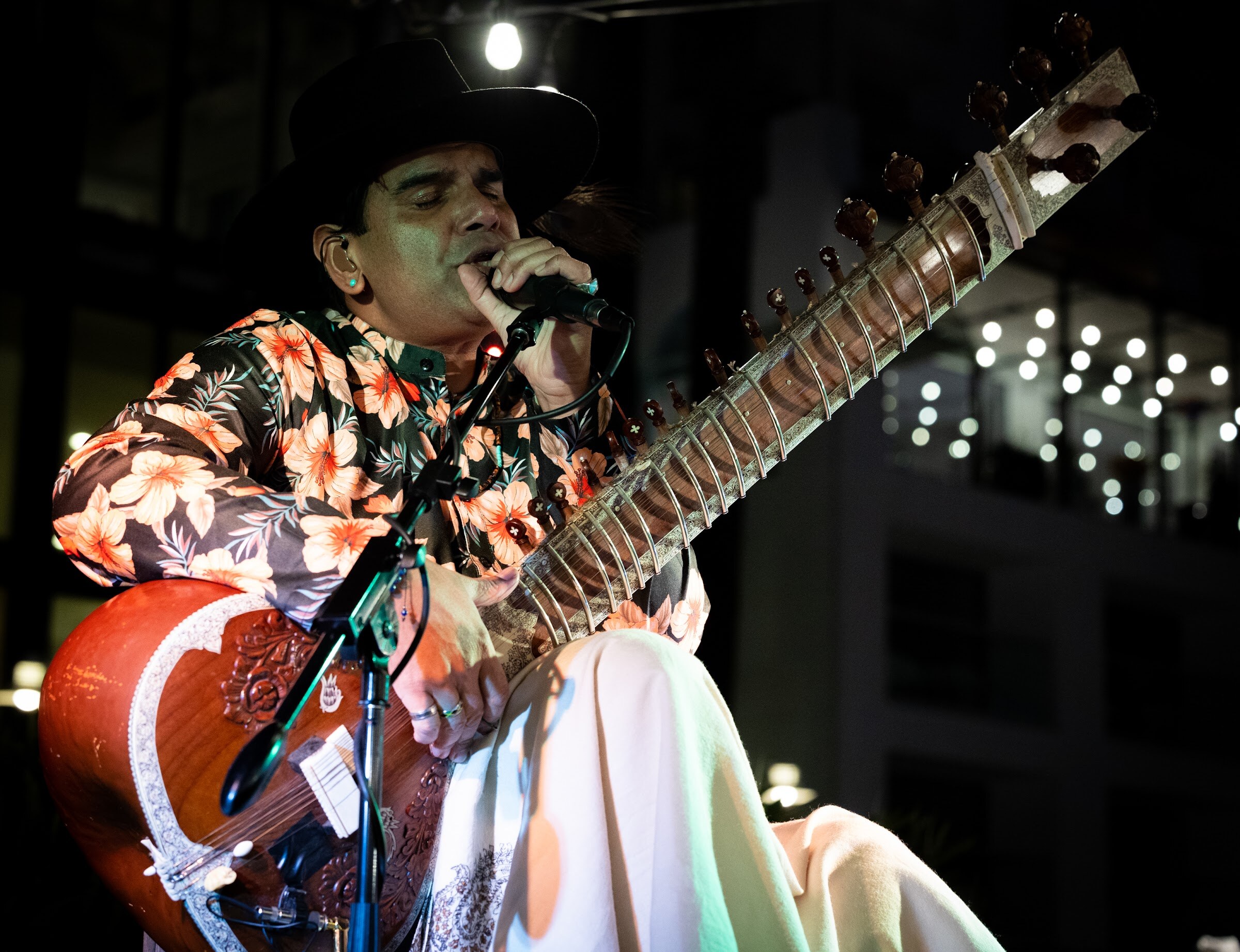 A performer in a black hat and orange floral shirt sings into a microphone while playing a sitar, with city lights behind him.