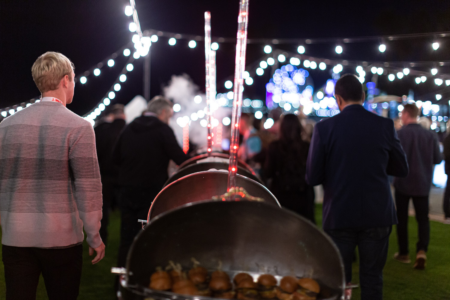 Two chafing dishes holding sliders in the foreground with attendees walking along a path lit by string lights.