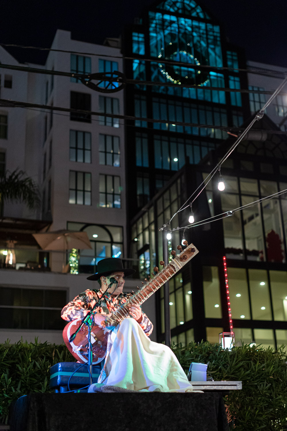 A performer sits cross-legged playing a sitar on a small platform with lit hotel buildings and string lights behind.