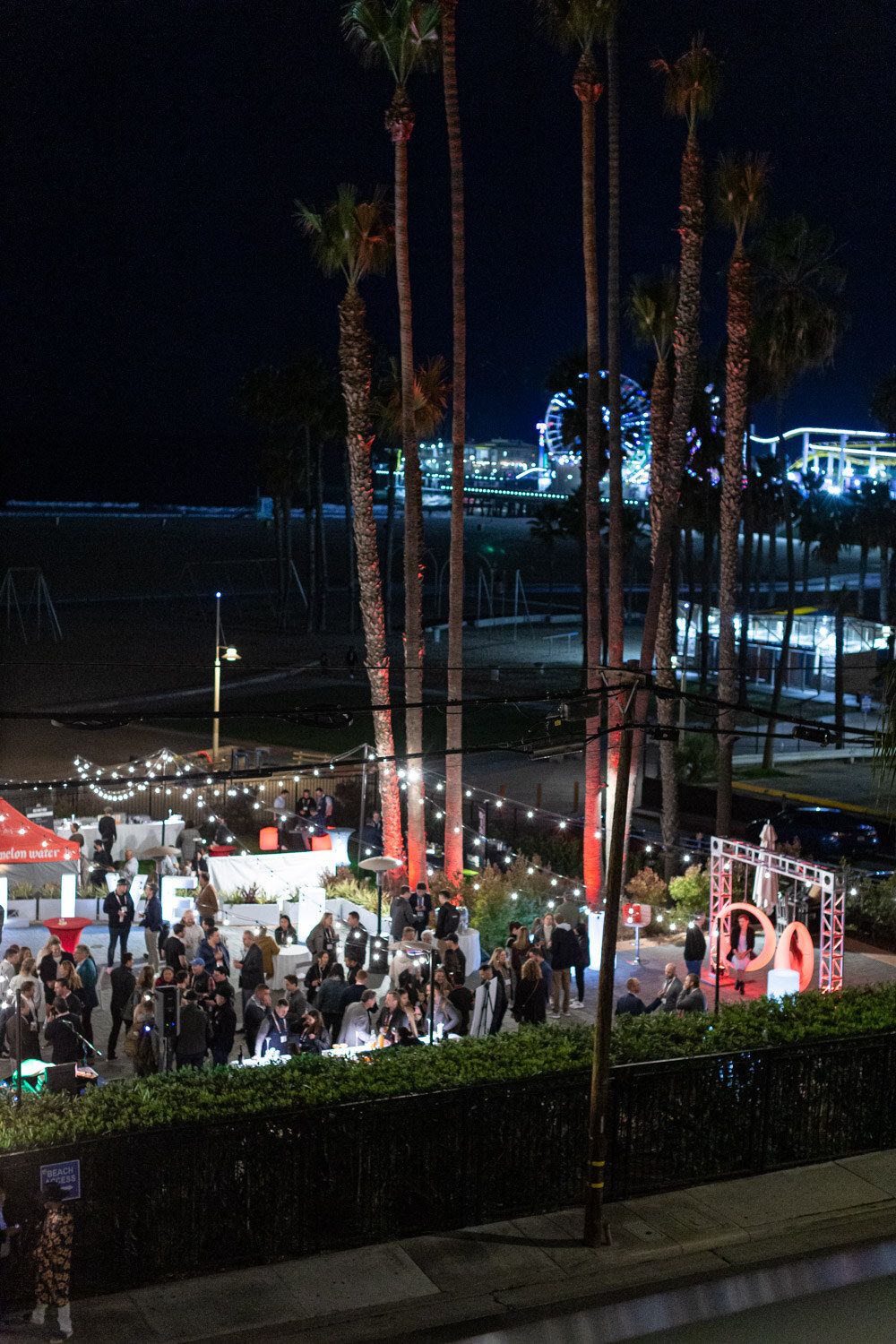 An elevated night view of a crowded lawn party with string lights, tents and palm trees, with the lights of the pier in the background.