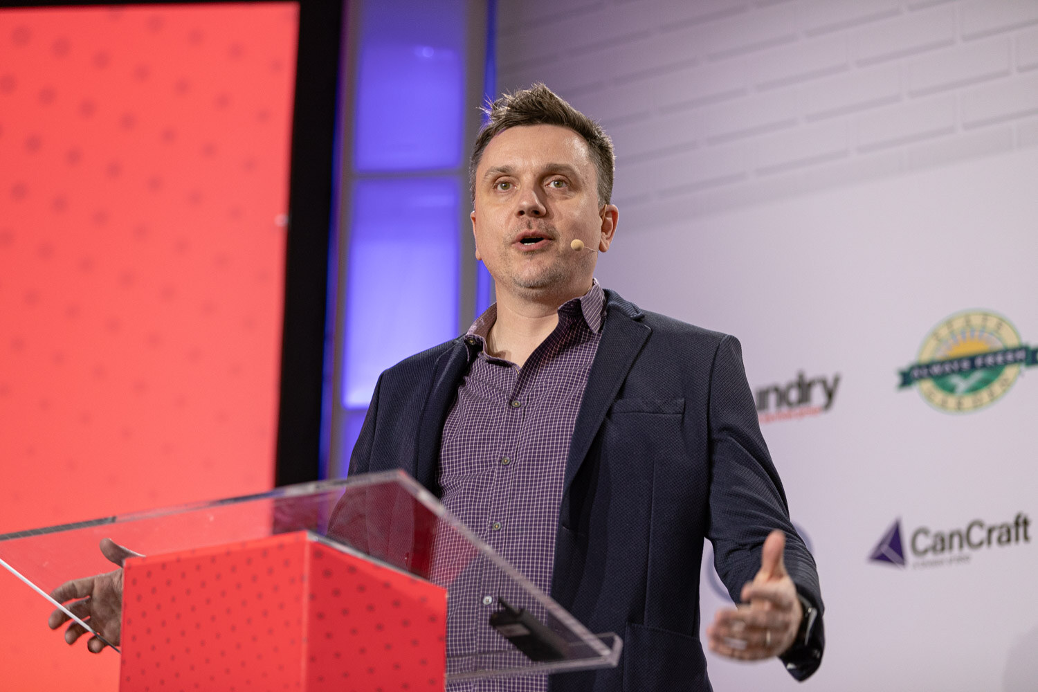 A man in a dark blazer and checked shirt speaks into a headset microphone at a clear podium in front of an orange-red screen.