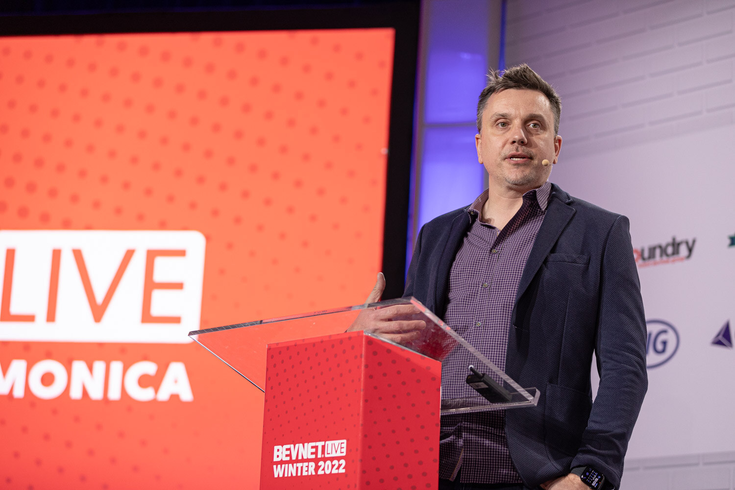 A man in a dark blazer speaks at a podium wearing a headset microphone, with a BevNET Live Winter 2022 podium front and part of the LIVE logo behind him.