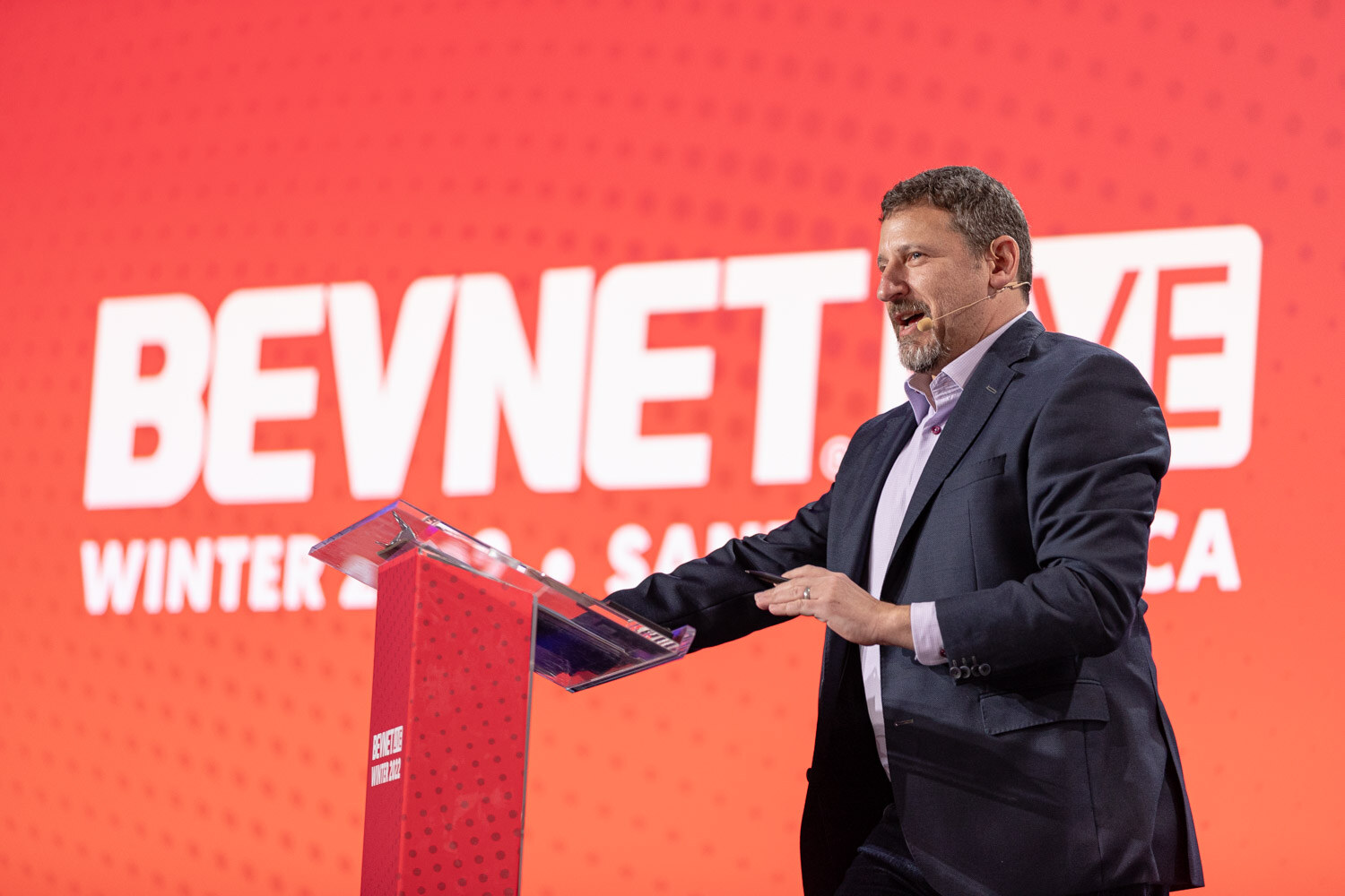 A bearded man in a navy jacket stands at a podium in front of a bright red screen reading BevNET Live Winter 2022 Santa Monica.