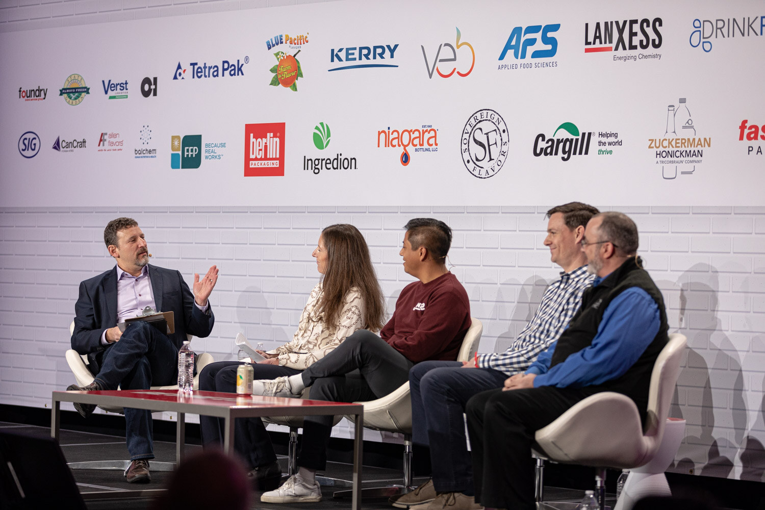 A wide shot of five people seated in white chairs around a low table on stage, in front of a wall of sponsor logos.