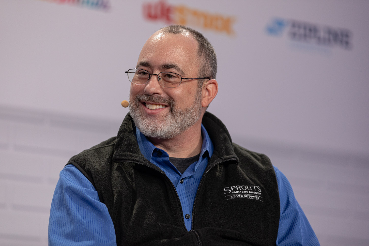 A bearded man in glasses, a blue shirt and a black fleece vest labeled Sprouts, wearing a headset microphone, smiles to his left.