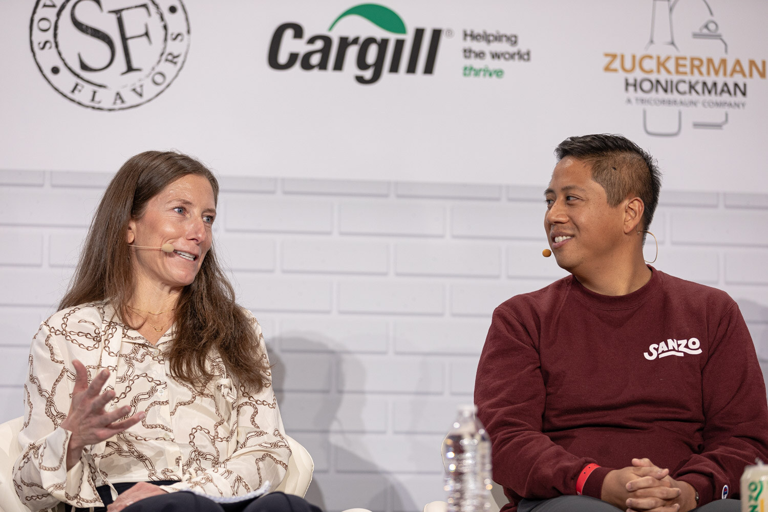 A woman in a patterned blouse and a man in a maroon sweatshirt with a Sanzo logo, both wearing headset microphones, sit side by side on stage.