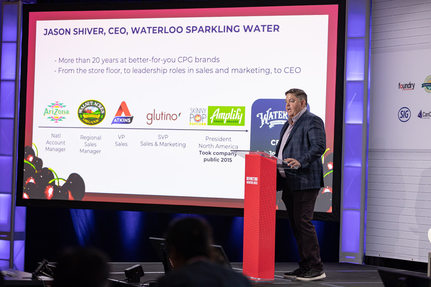 A man stands at a red podium beside a large screen showing a career timeline with logos and a Waterloo Sparkling Water CEO title.