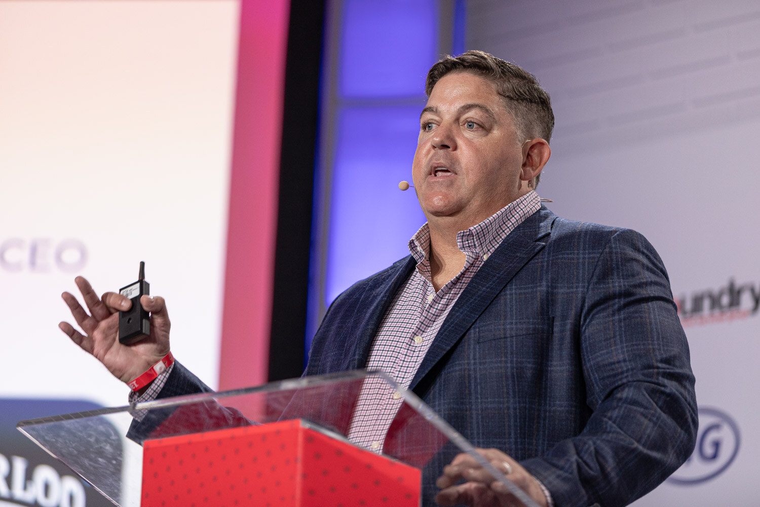 A man in a plaid blazer holds a presentation clicker and speaks at a clear podium on a lit stage.