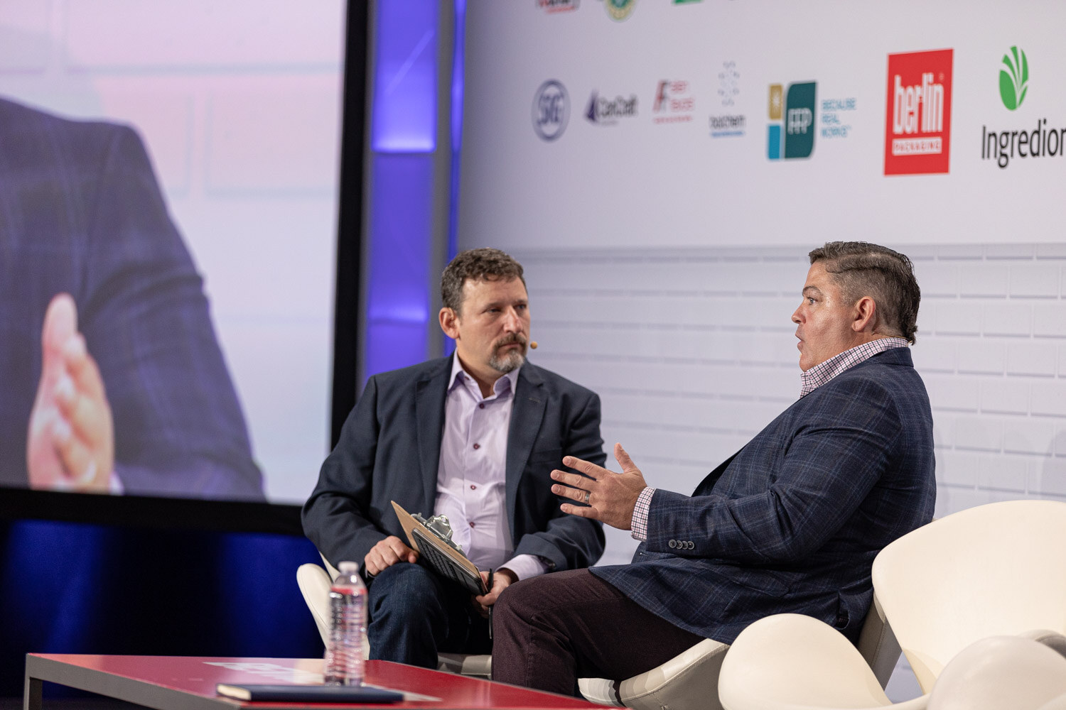 Two men sit on white chairs on stage, the moderator on the left holding a clipboard and the speaker on the right gesturing.