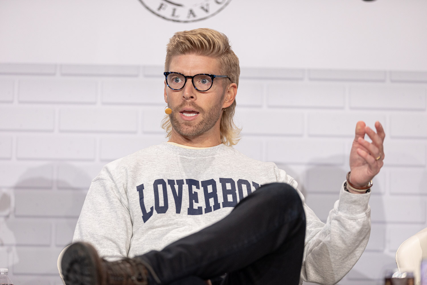 A blond man in dark glasses and a gray Loverboy sweatshirt, wearing a headset microphone, gestures with one hand while seated with his leg crossed.
