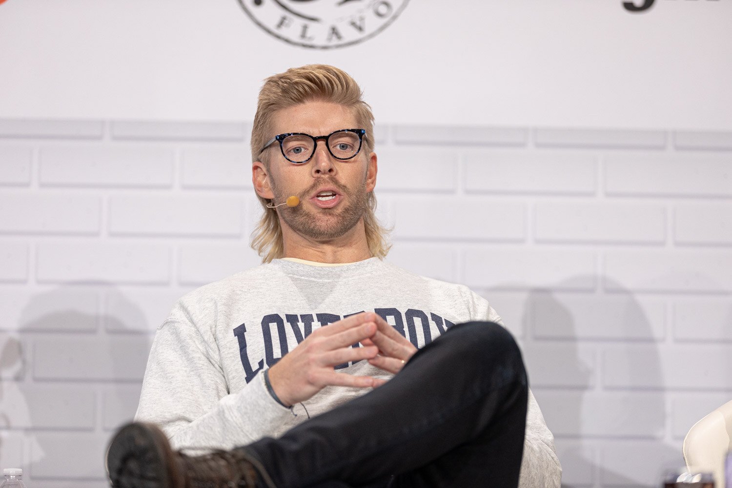 A blond man in dark-framed glasses and a gray sweatshirt with partly visible lettering presses his fingertips together while speaking.