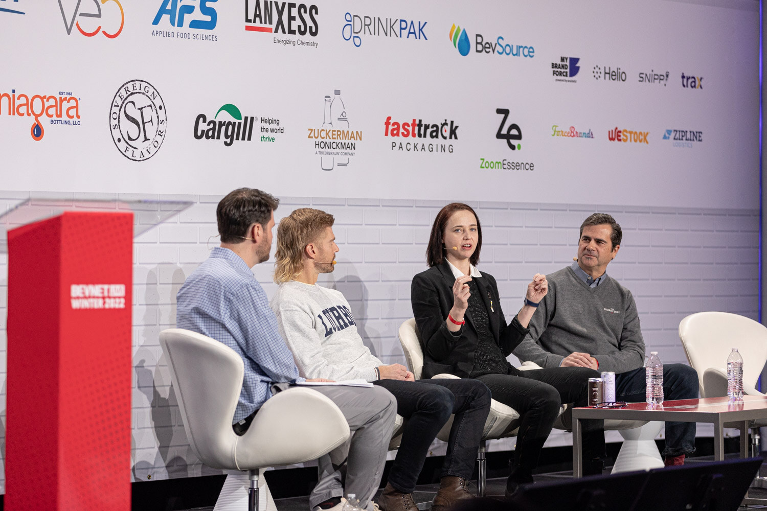 Four people sit on white chairs on a stage beside a red podium in front of a sponsor wall, with one woman gesturing as she speaks.