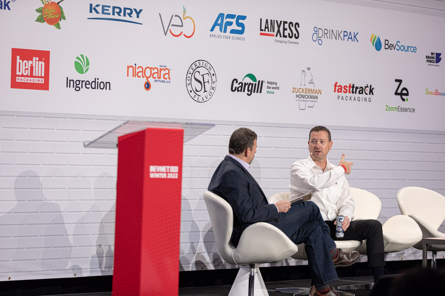 Two men sit in white chairs on stage beside a red podium in front of a wall of sponsor logos including Kerry, AFS and Lanxess.