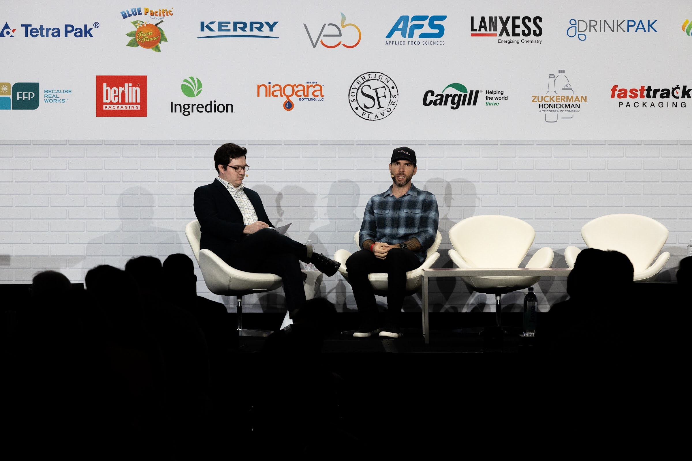 Two men sit on white swan chairs on the stage in front of a BevNET Live sponsor wall, with a darkened audience in front.
