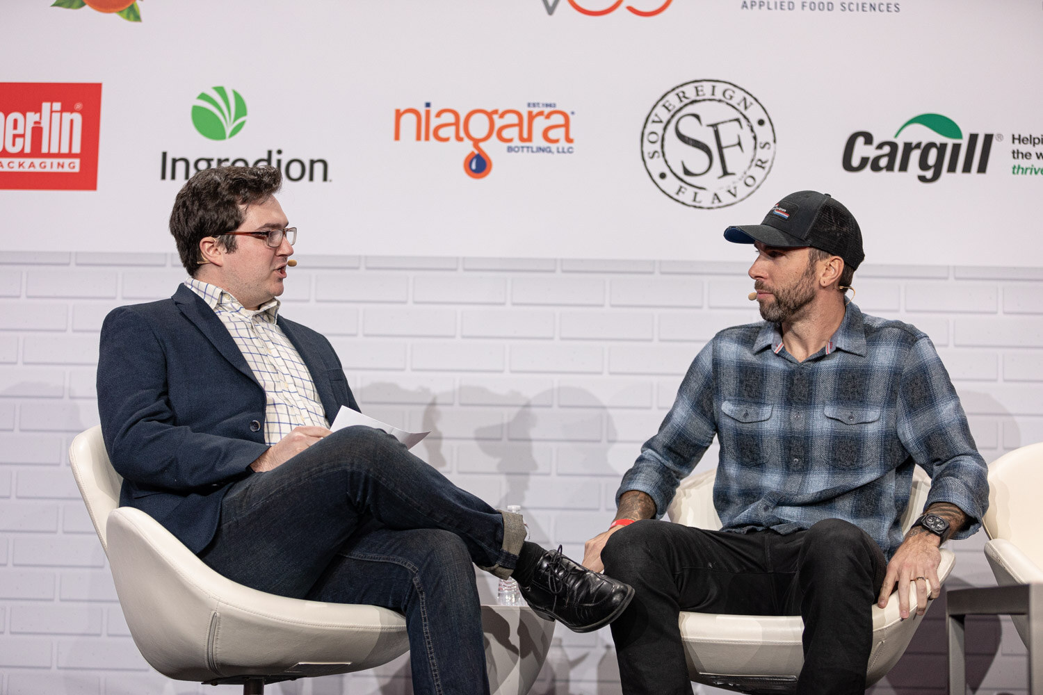 A man in a navy blazer holding papers and a bearded man in a plaid shirt and cap sit in white chairs in front of a sponsor wall.