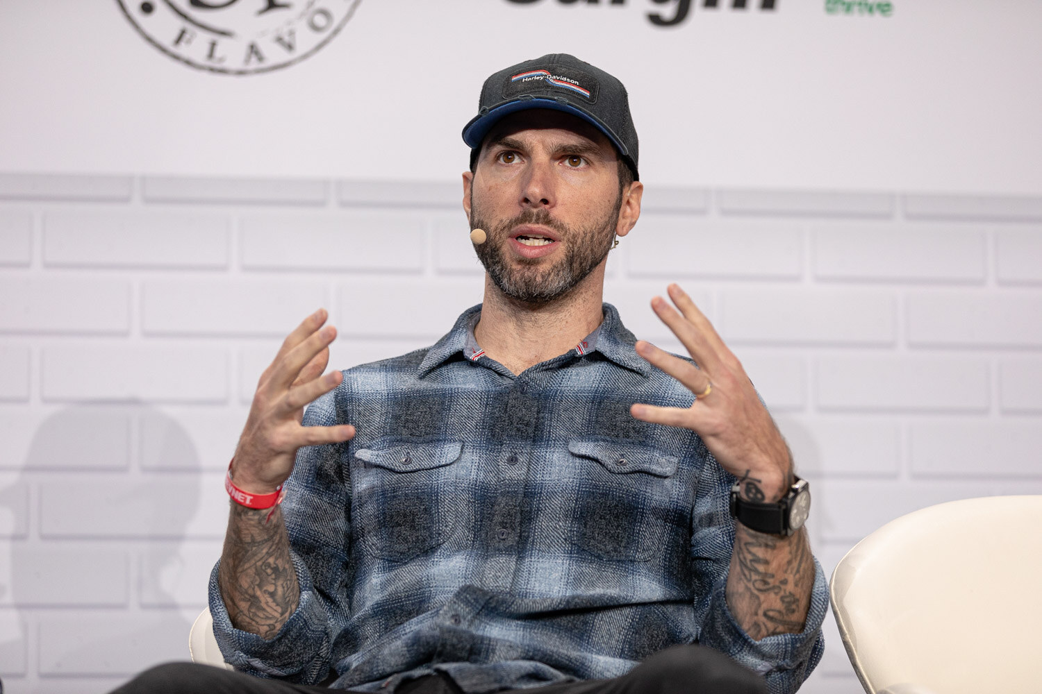 A tattooed man in a ball cap and blue plaid shirt holds both hands up as he speaks, wearing a headset microphone.