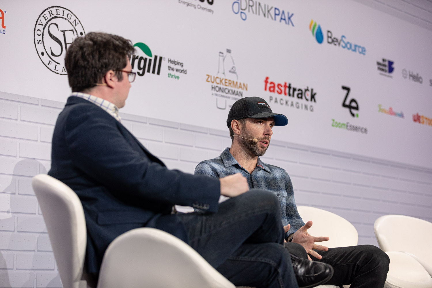 Two men sit on white chairs, the one in the foreground in a navy blazer and the other in a plaid shirt and baseball cap, in front of a sponsor wall.