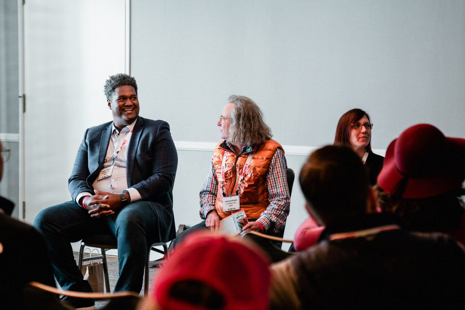 Three seated panelists in a hotel breakout room, with a man in a navy blazer smiling at a long-haired man in an orange vest, and attendees in red caps in the foreground.
