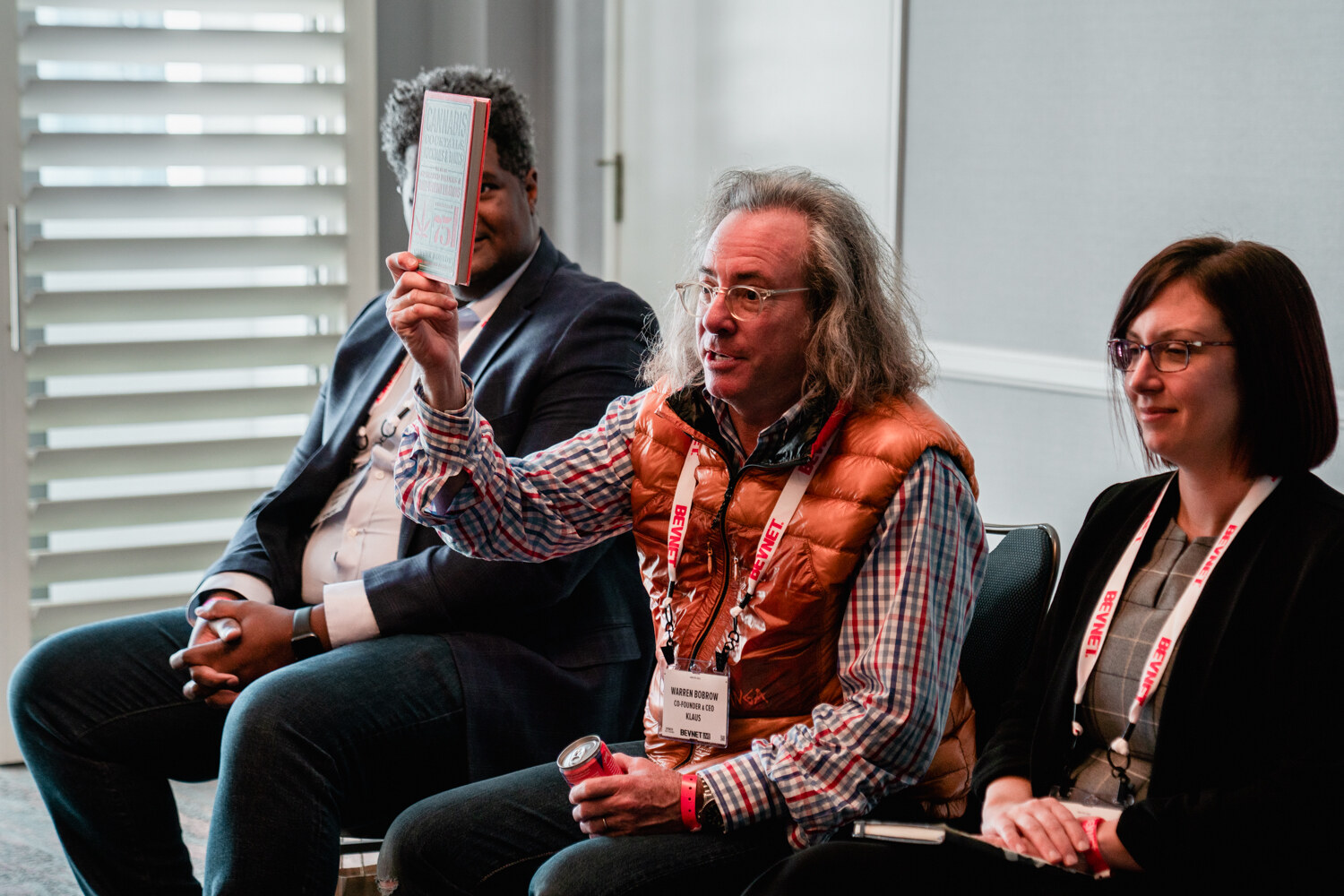 A long-haired man in an orange vest raises a book above his head while seated between two other panelists in a breakout room.