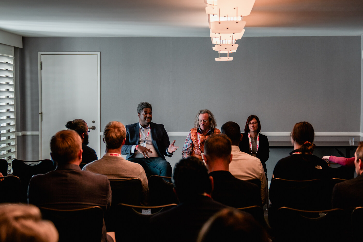 A dim breakout room with rows of attendees seen from behind facing three seated panelists beneath a chandelier-style light.