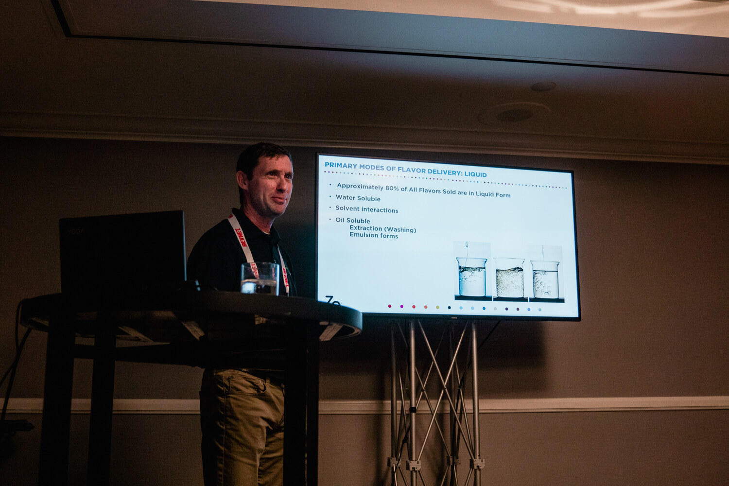 A man stands at a lectern beside a screen titled Primary Modes of Flavor Delivery: Liquid in a dimly lit room.
