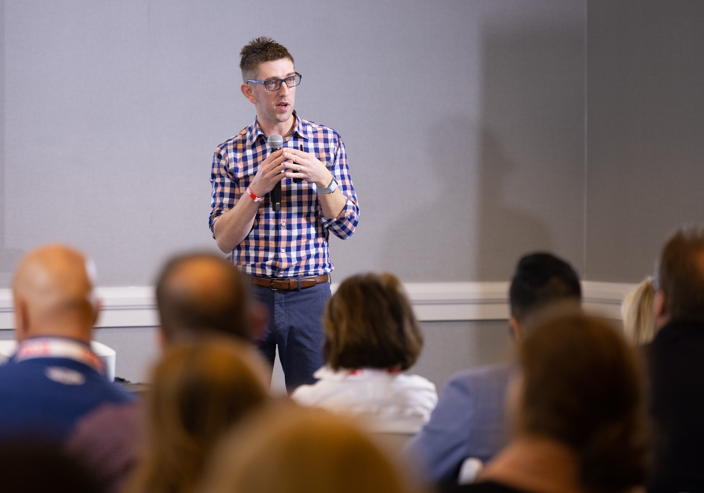 A man in a blue and white checked shirt holds a microphone in front of a gray wall while attendees sit in the foreground.