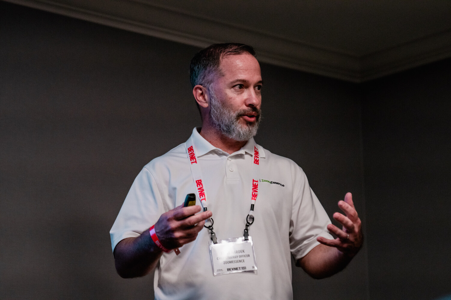 A bearded man in a white polo with a BevNET lanyard and ZoomEssence badge holds a clicker and gestures with one hand in a dim room.
