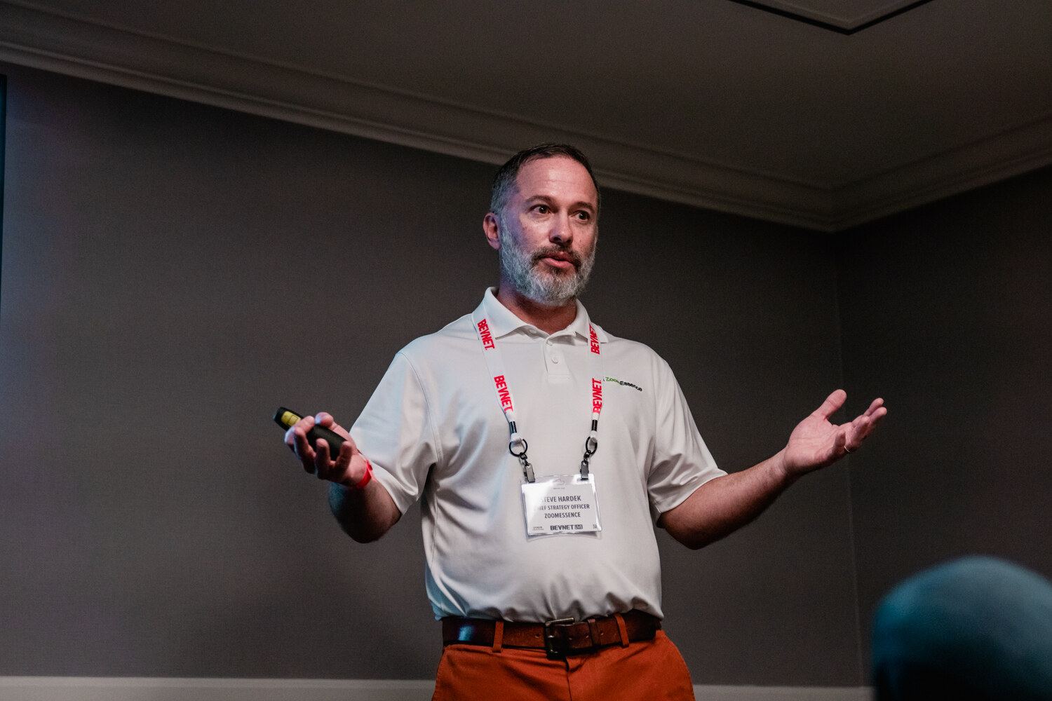A bearded man in a white polo and orange pants holds a clicker in one hand and spreads both arms in front of a gray wall.