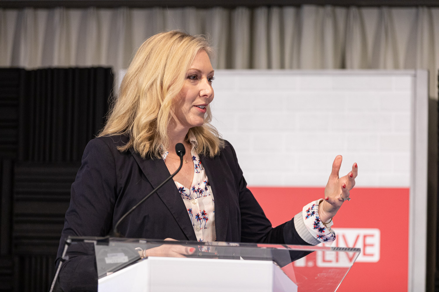A blond woman in a navy blazer and floral blouse speaks into a microphone at a podium with a BevNET Live sign.