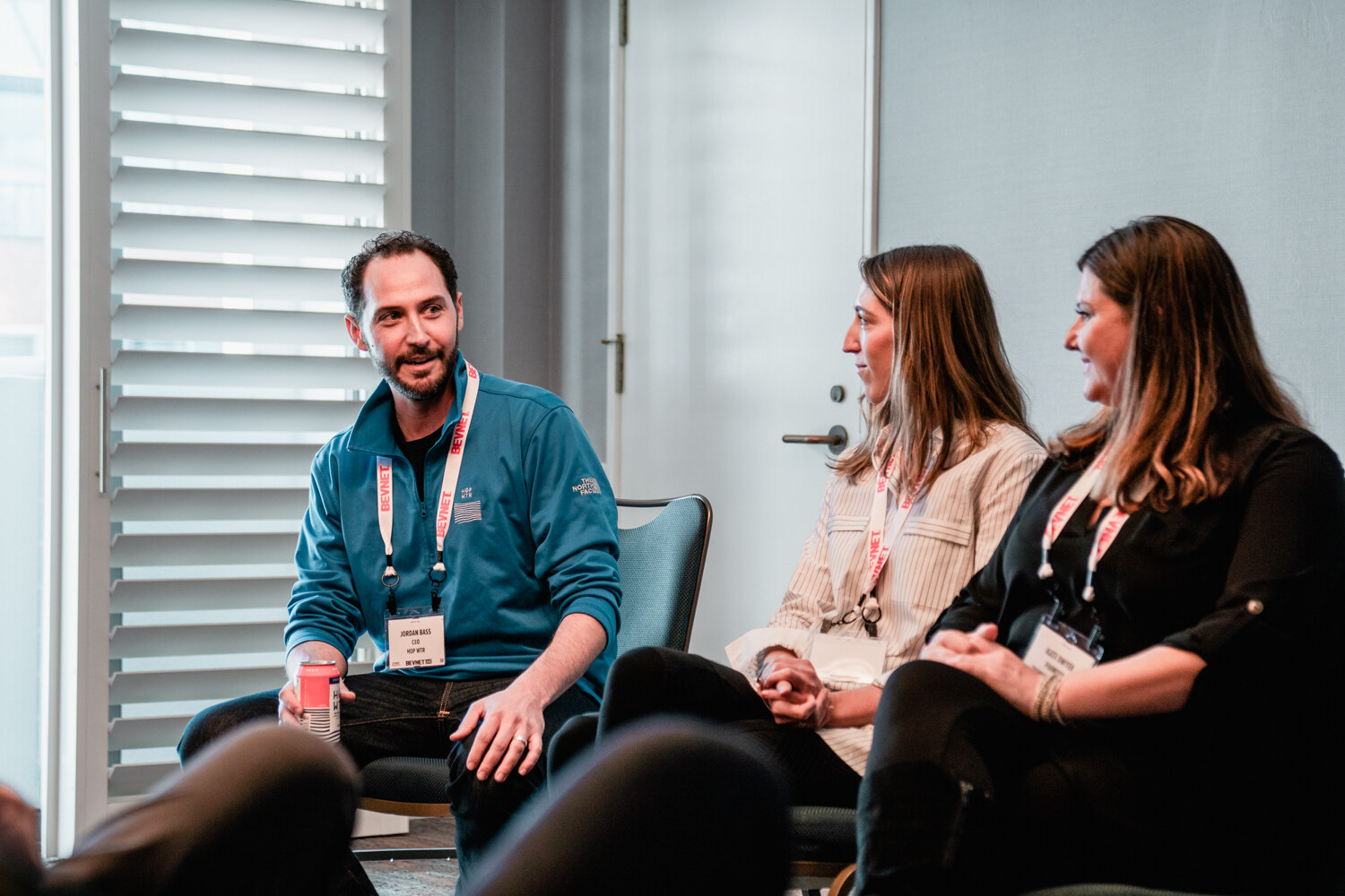 A man in a teal fleece and two women, all wearing BevNET Live badges, sit in chairs beside a window with blinds.