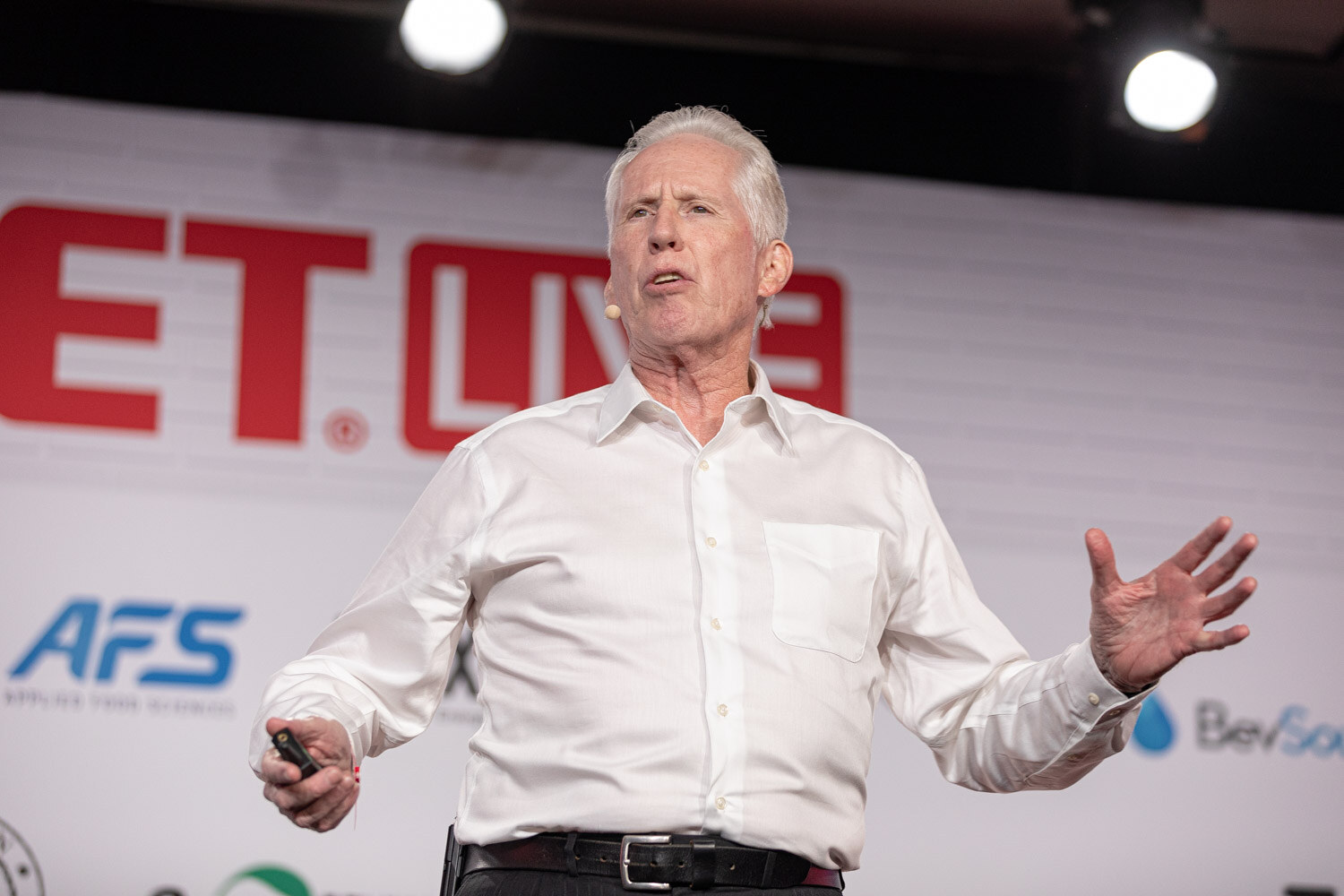 A white-haired man in a white shirt wearing a headset microphone holds a clicker and spreads both hands in front of a BevNET Live sign.
