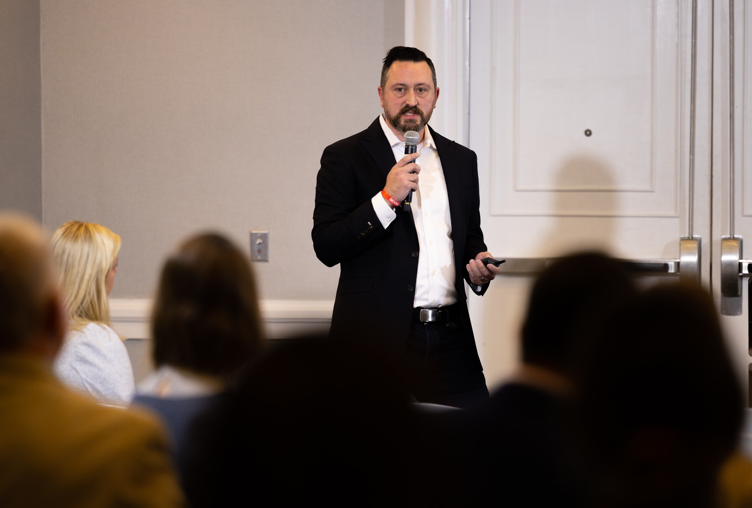 A bearded man in a dark blazer holds a microphone while speaking to seated attendees seen from behind in a hotel meeting room.