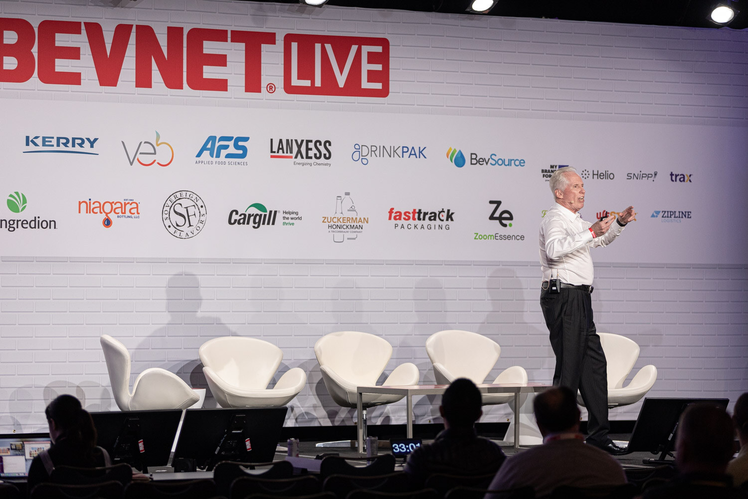A man stands alone on a stage in front of a BevNET Live wall of sponsor logos, with white swan chairs behind him and the audience in silhouette.