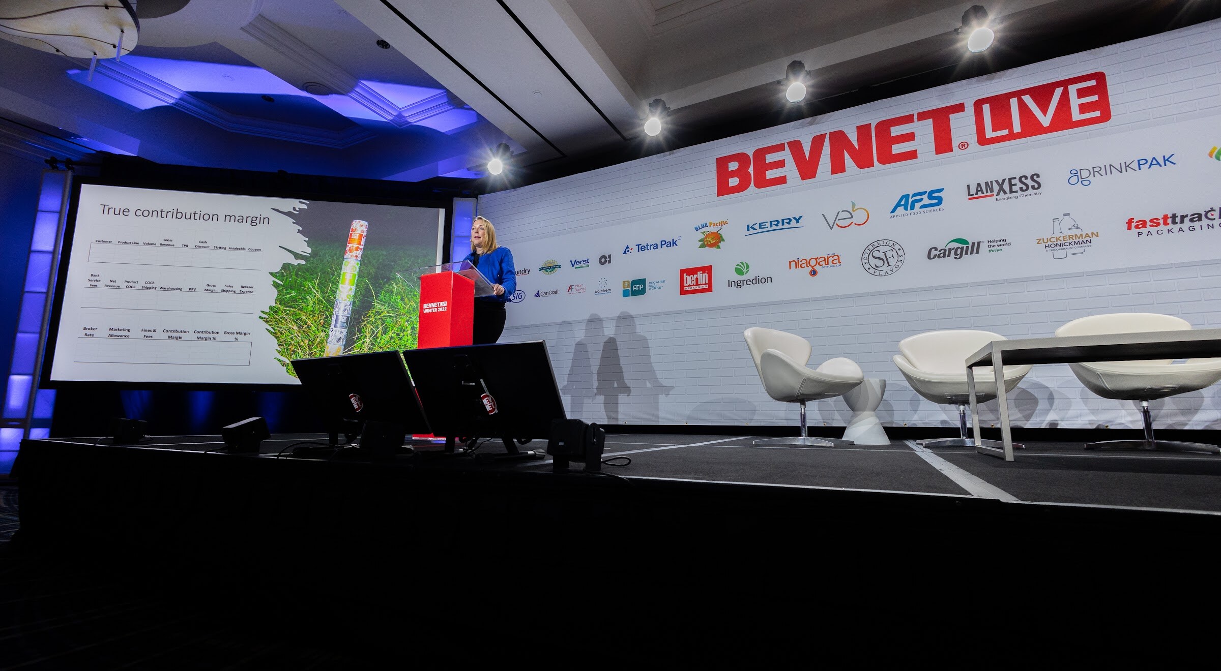 A wide shot of the main stage showing a woman at a red podium beside a screen reading True contribution margin, with BevNET Live and sponsor logos on the wall.