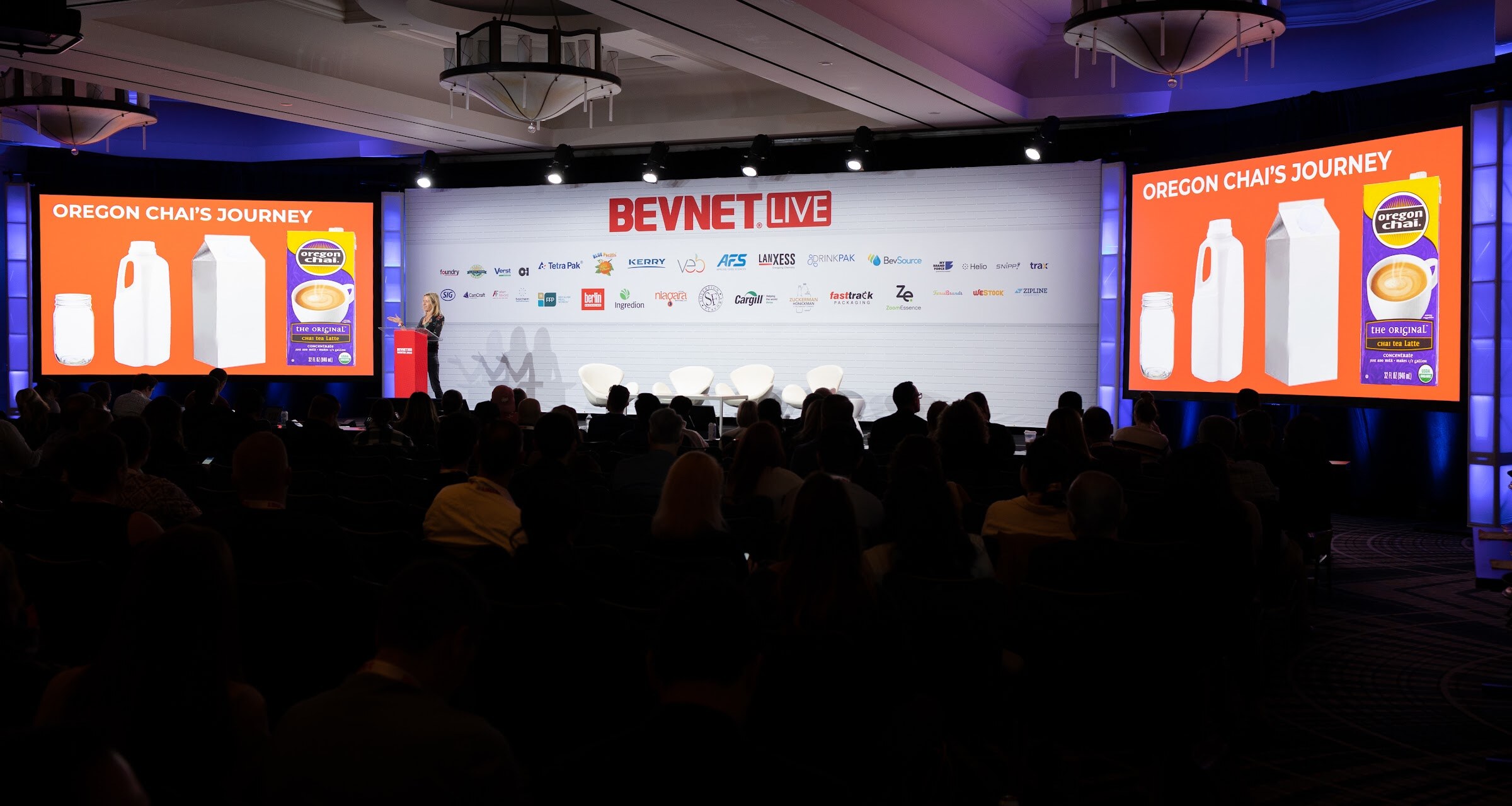 A wide view of the dark ballroom with a full audience facing a stage and two large orange screens reading Oregon Chai's Journey.