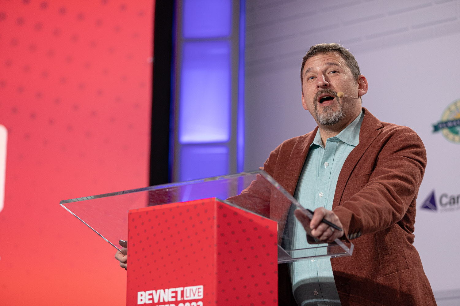 A bearded man in a rust-colored blazer and teal shirt speaks into a headset microphone and points from behind a clear podium in front of an orange screen.