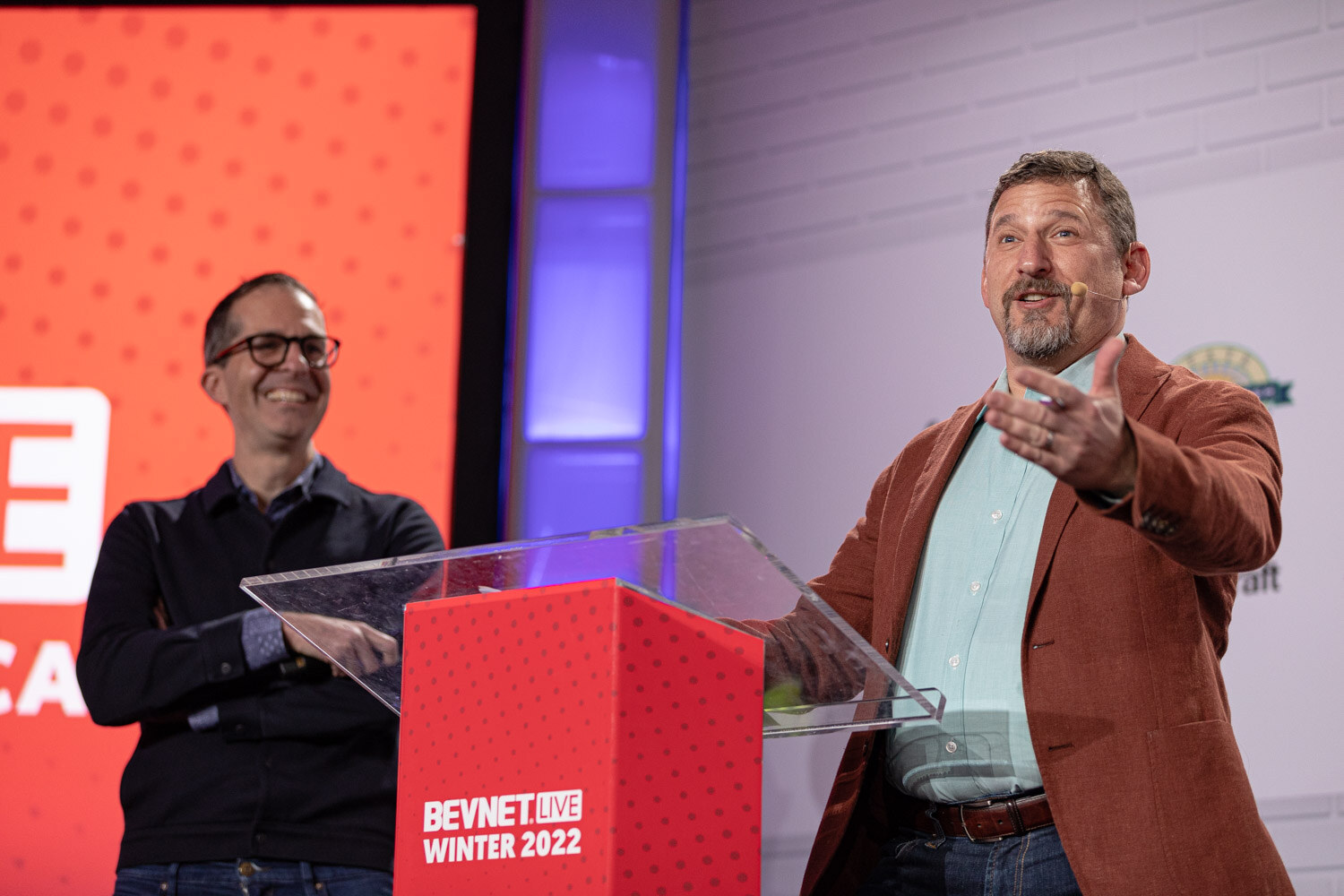 A man in a rust blazer with a headset microphone gestures from a podium while a man in dark glasses and a black sweater smiles with arms crossed behind him.