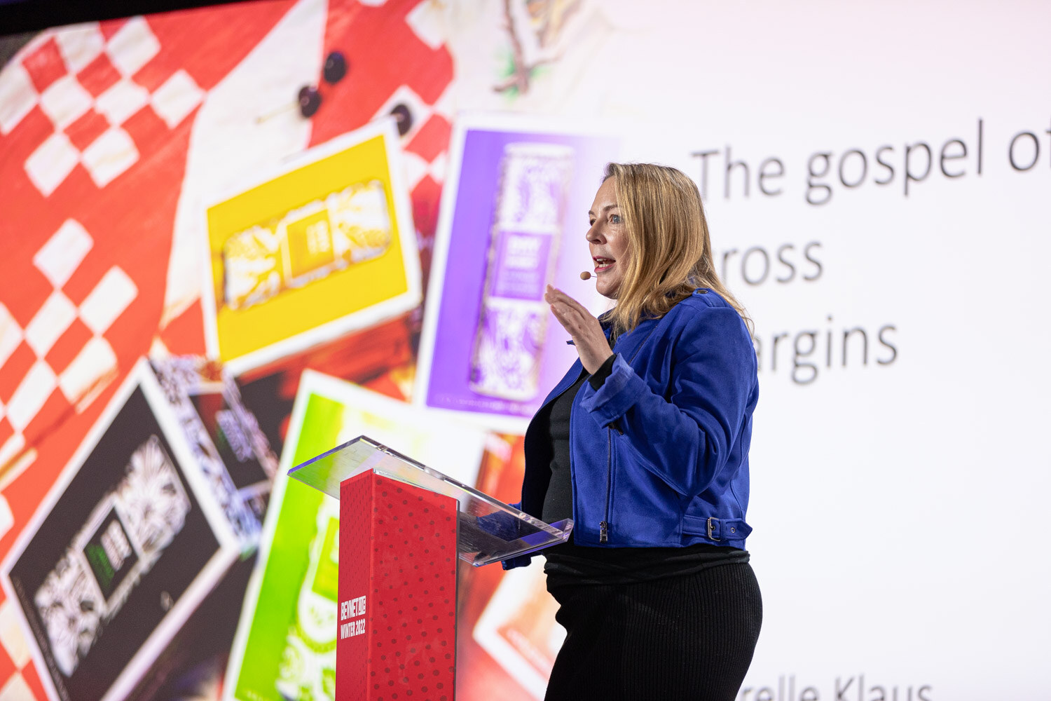 A woman in a blue jacket speaks from a podium in front of a colorful slide with a checkerboard background and the text The gospel of gross margins.